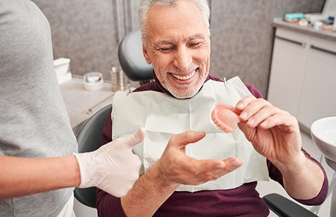 Man in dental chair smiling holding dentures with dentist giving thumbs up